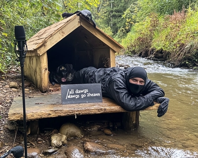 A masked man in all black laying on top of a doghouse with a sign that says "All Dawgs Go 2Heaven". It is next to a stream and in the background there is a microphone and bacon pancakes