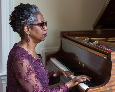 A van Gogh inspired portrait of an African-American woman in her 40s with very short, gray and black natural hair wearing wide cat eye framed glasses and wearing a plum color dress while playing an old  upright brown piano in her living room