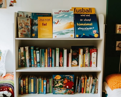 a child's room with a poster on the wall. There is no bookshel in the room, please show a large rom with bed, writing desk, carpet etc. The following text describes what to be seen in the poster: a white children's bookshelf with three tiers. This bookshelf in the poster: top shelf displays larger picture books standing upright, including:
"Fußball" (a soccer-themed book)
"Wind" by Kenneth Grahame
"Der Schlumpf der vom Himmel fiel" (A Smurf book with yellow cover)
"Junior" series book
"Alles Erlaubt?"
The middle shelf contains a row of smaller books packed tightly together, with some DVD cases visible on the right side.
The bottom shelf holds what appears to be board games and other children's materials."