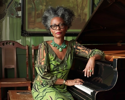 a Monet inspired picture of an African-American woman around 50 years of age with short, gray and black coily Afro and wearing wide black frame cat eyeglasses and also wearing a chartreuse 60s style dress while sitting on her piano bench with her hands fold in her lap in front  of her old vintage upright, black Piano